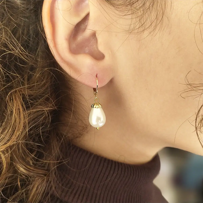 Lucrezia earrings with 10x14 mm. shell pearls with opalescent finish and gold-plated caps holding the pearls up to the clasp on a ear model shown at a close distance.