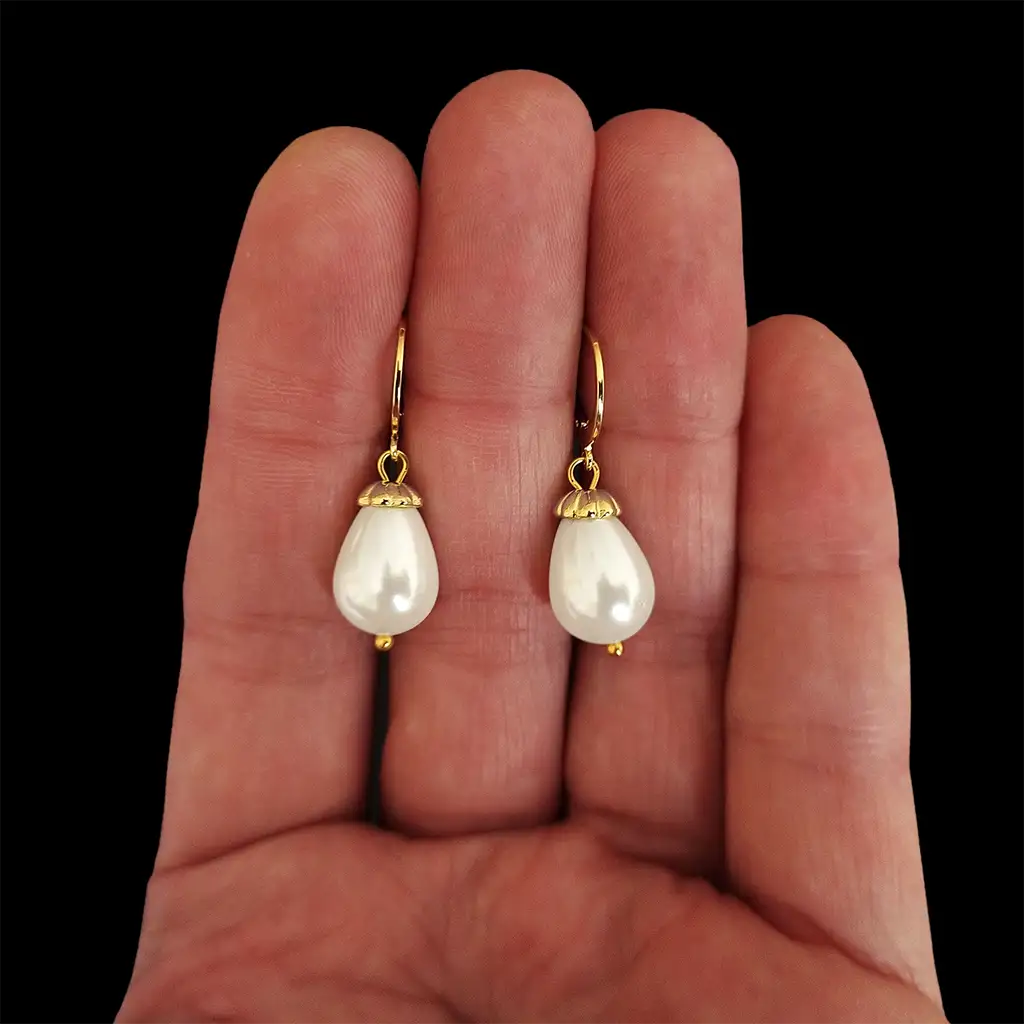 Close up view of the Lucrezia earrings with 10x14 mm. shell pearls with opalescent finish and gold-plated caps holding the pearls up to the clasp shown on the fingers of one hand.